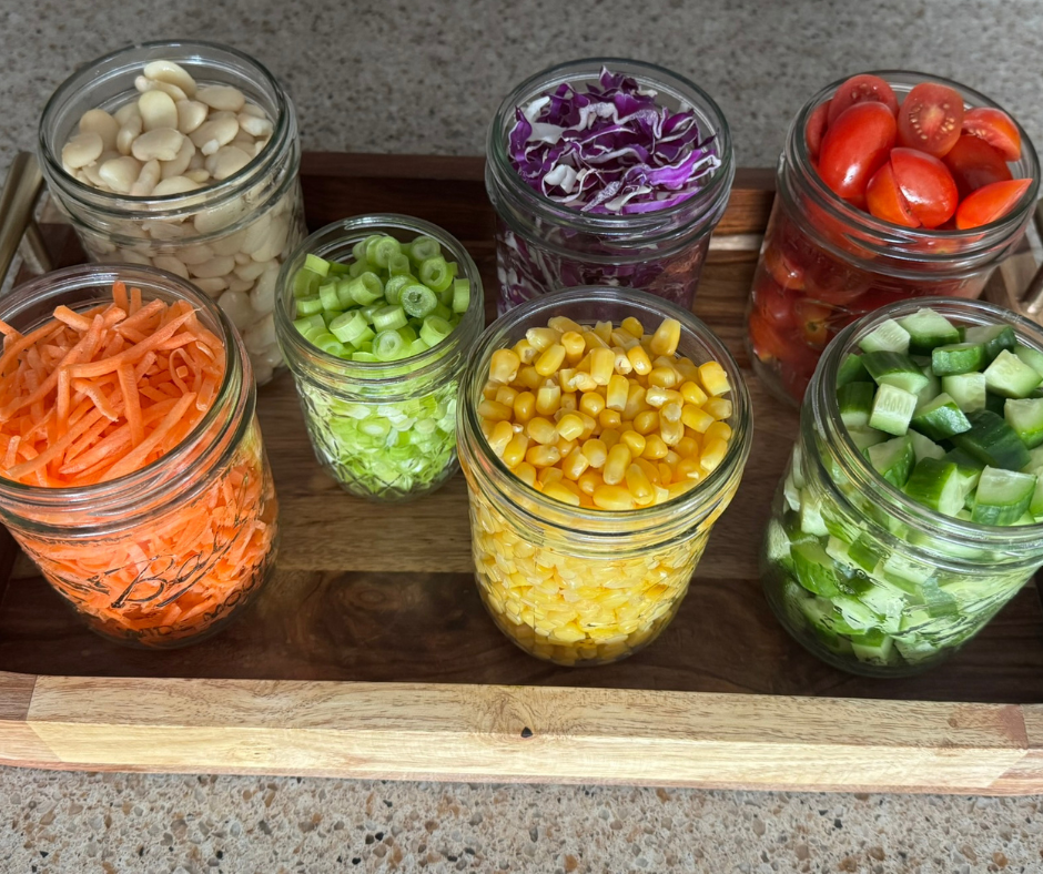 Prepped Salad of the Week ingredients stored in glass jars on a tray, including butter beans, corn, chopped vegetables, greens, and herbs ready for quick assembly