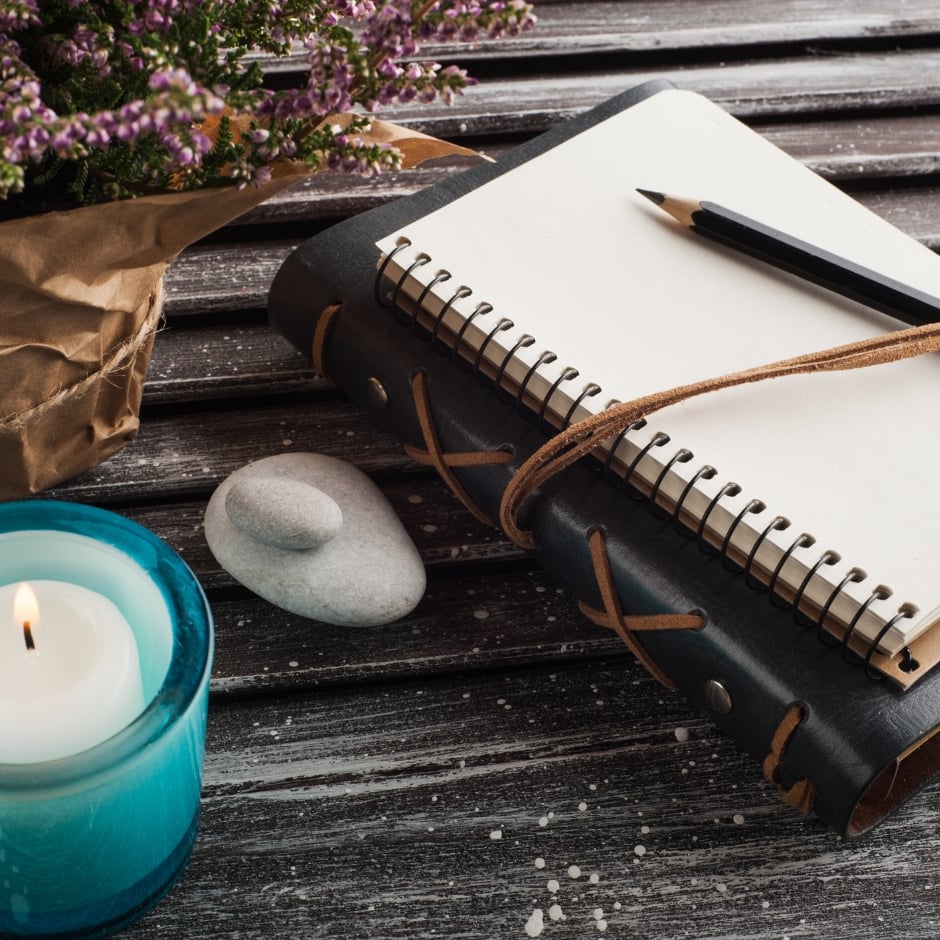 Calm, holistic wellness scene featuring a spiral-bound leather journal, a lit candle in a soft blue glass holder, a small stone, and a potted plant with purple flowers, all arranged on a rustic wooden surface. The scene evokes a sense of peaceful mindfulness, ideal for supporting themes of intentional, natural wellness practices.